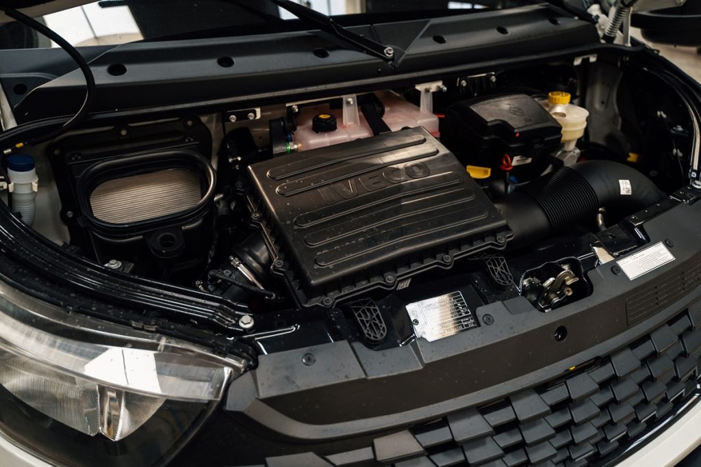 Zdjęcie przedstawia otwartą komorę silnika pojazdu Iveco z dużą, czarną plastikową pokrywą z napisem „IVECO”, widocznym różowym zbiornikiem płynu chłodniczego oraz innymi elementami układu napędowego i osprzętu