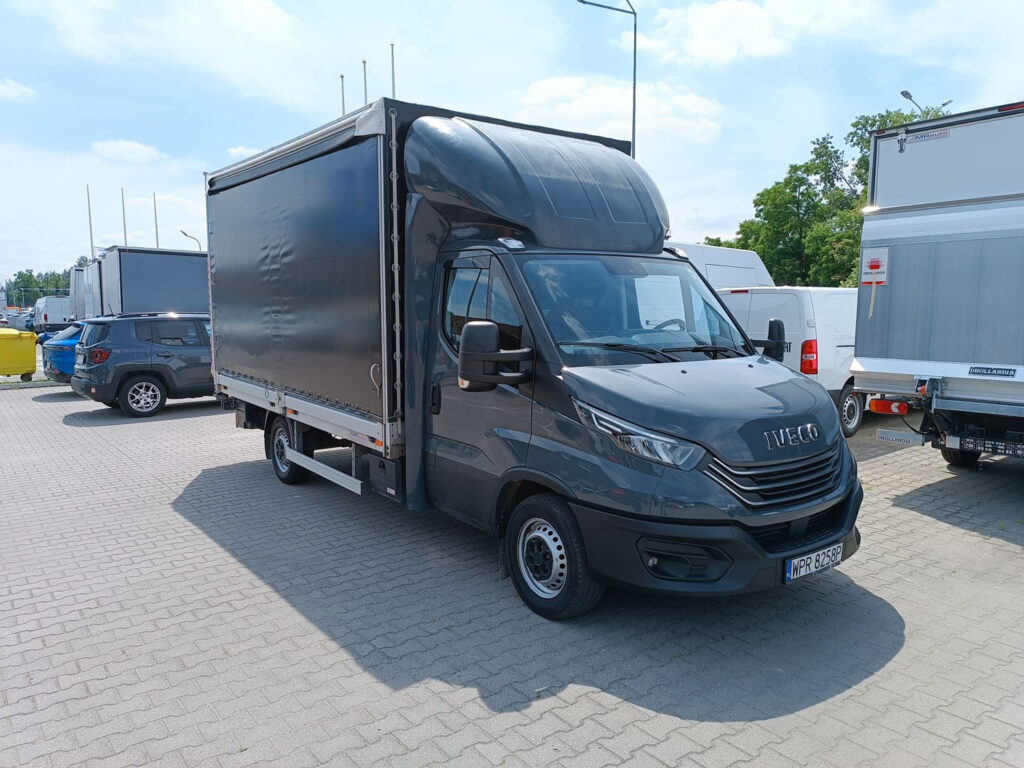 **Alt tekst:** Ciemnoszare Iveco Daily z zabudową plandekową zaparkowane na placu sprzedażowym Uni-Truck, otoczone innymi samochodami dostawczymi.