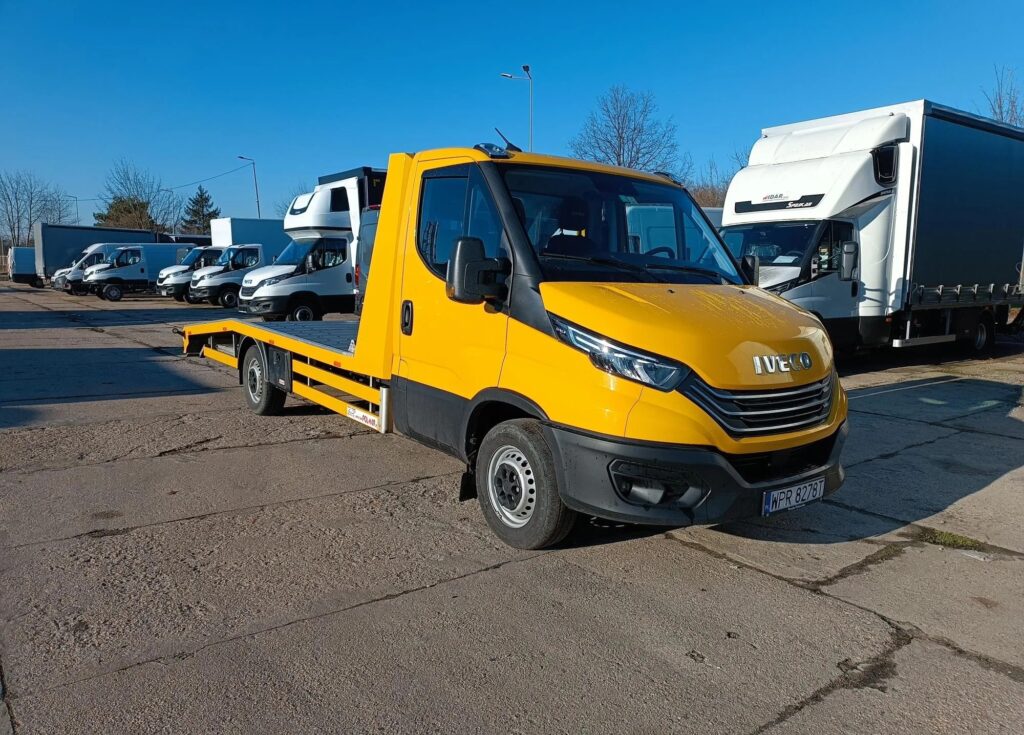 Żółta autolaweta Iveco Daily zaparkowana na placu przed innymi samochodami użytkowymi – przykład uniwersalnego rozwiązania do transportu pojazdów.